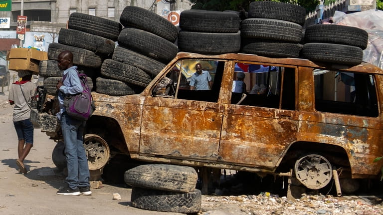 People walk past tires and a burned-out vehicle used to...