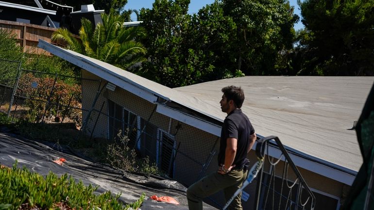 A reporter stands near a home that collapsed due to...